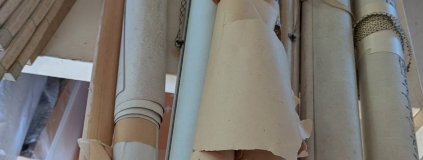 神奈川県横浜市西区の空き家片付け中に見つかった掛軸の束。和紙に包まれた古い日本画・書道作品の掛軸が机の上に並び、経年による変色や包み紙の破れが見られる様子。