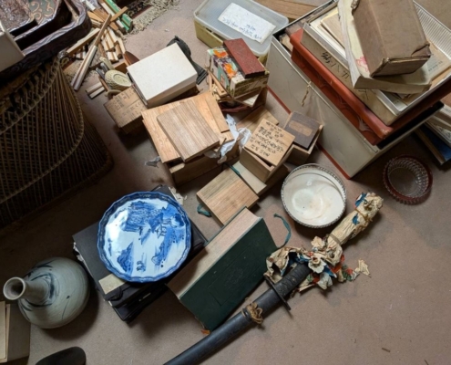 空き家となった実家の片付け中に見つかった明治時代の骨董品。木箱入りの書道具、和本、染付の皿、陶器、刀剣などが床に広げられている様子。